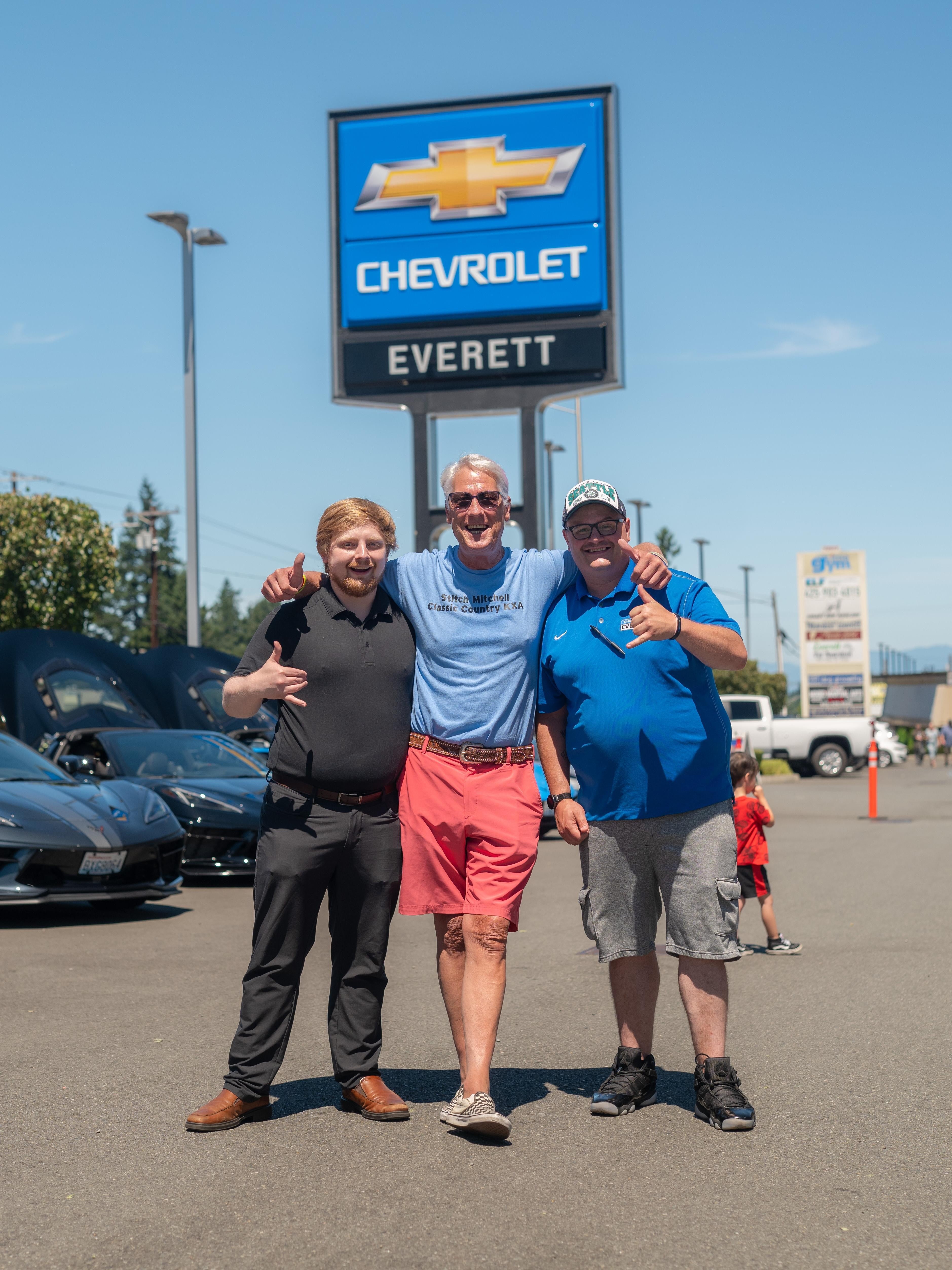 Three men pose in front of a Chevrolet dealership, showcasing a variety of cars in the background.